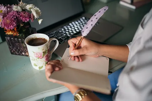 コーヒーを飲みながら羽根ペンでノートに書き物をする手元